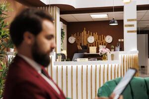 Hotel reception and lounge area with tourists waiting for check in process, man preparing for business meeting at conference table. Front desk lobby with white collar worker, work trip. photo