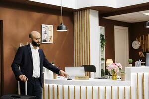 Businessman at hotel front desk using service bell to ring for concierge assistance, preparing for room registration. Entrepreneur checking in at resort, corporate business conference. photo