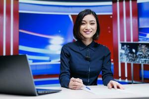 Asian presenter on daily newscast in newsroom, talking about latest international events on live broadcast. Woman reporter creating television content with media outlets headlines. photo