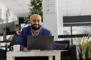 sonriente árabe empresario trabajando en ordenador portátil en negocio oficina mientras sentado en sofá. comienzo arriba empresa ejecutivo gerente analizando exitoso proyecto ventas en portátil computadora en trabajo colaborativo espacio foto