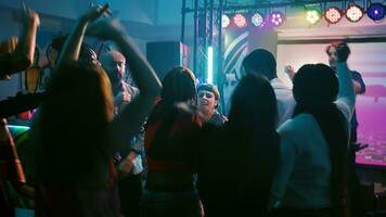 Young adults dancing on electronic music, having fun together with DJ on stage. Cheerful men and women jumping on dance floor under disco lights, enjoying dance party. Handheld shot. photo