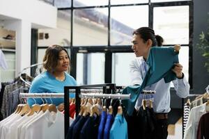 African american customer discussing clothes fabric, looking at racks full with fashionable merchandise in clothing store. Stylish woman choosing to buy casual wear in modern boutique photo