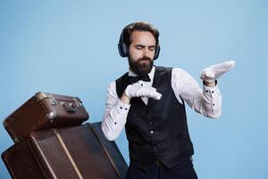 Hotel staff member dancing on camera, having fun with music on headphones while he wears elegant clothes and gloves. Young man worker with audio headset feeling confident or funky. photo