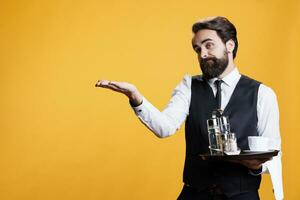 Elegant butler making way for clients to come at restaurant, helping them with seats at table before serving food. Professional catering employee providing luxury services, holds tray. photo