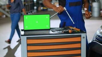 Green screen laptop placed on working bench in busy garage next to professional tools while repairmen fix cars in blurry background. Chroma key device in auto repair shop, panning shot photo