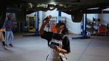 Qualified engineer underneath suspended car in repair shop, using work light to check for damages during maintenance. Expert using professional tool and tablet to examine vehicle on overhead lift photo