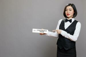 Asian woman receptionist holding conference room directional tabletop signage and pointing with finger portrait. Hotel employee in uniform showing boardroom steel sign and looking at camera photo