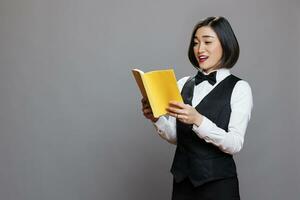 Focused smiling asian woman receptionist holding book with yellow cover and reading. Cafeteria young waitress wearing uniform enjoying novel paperback while learning literature photo