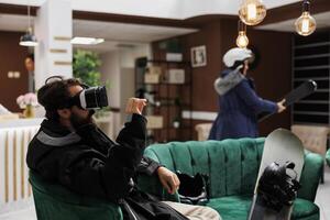 Young male traveler in hotel lobby during wintertime embracing technology by using vr gadget. Visitor exploring internet in lounge area. Snowboarding gear and helmet suggests ski mountain resort. photo