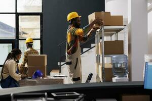African american warehouse managers team picking buyer order and choosing cardboard box for packing. Woman checking goods list on laptop and man standing on ladder while taking parcel from shelf photo