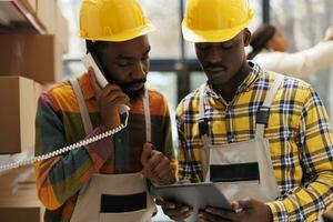 African american storehouse employees coordinating delivery schedule with logistics manager on landline telephone. Warehouse operator talking on phone and looking at digital tablet screen photo