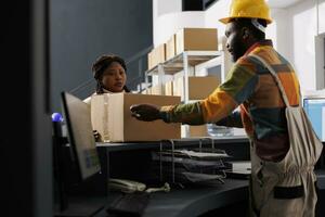 Postal service worker managing parcel receiving and registration in warehouse. African american woman courier putting cardboard box on counter desk in shipment company storehouse photo