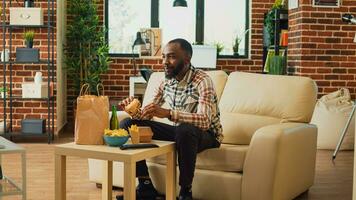 Cheerful man taking bite of hamburger in living room, eating fast food from takeaway delivery order. Happy adult watching television at home and serving burger with fries and alcohol. photo