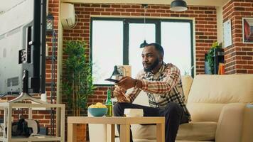 African american guy eating noodles in living room, sitting at television and watching movie. Young man serving asian food with chopsticks, ordering dinner from takeaway restaurant. photo