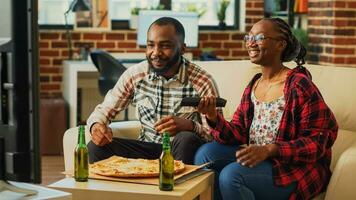 Cheerful man and woman eating slices of pizza at home, being relaxed together watching movie on television. Young people in relationship enjoying delivery meal and beer bottles. photo