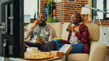 African american people watching film on tv together, drinking beer and eating chips on sofa. Modern couple feeling relaxed with film on television and multiple types of fast food from delivery. photo