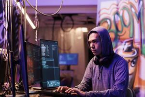 Asian hacker cracking password to get illegal access to database confidential information. Young man in hood infecting victim computer with internet virus in abandoned warehouse photo