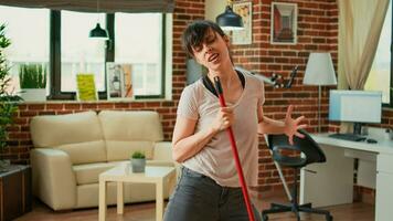 Silly woman mopping floors with washing solution and dancing in living room, spring cleaning. Young cheerful housewife using mop to sweep dirt and clean mess, listening to music. photo