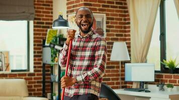 Modern happy boyfriend having fun with music while he uses mop to wash floors, mopping in apartment. Young male adult dancing and singing in living room, spring cleaning enjoyment. photo