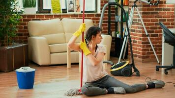 Sleepy housewife sitting on apartment floors with mop and bucket, feeling proud about finishing spring cleaning session. Young woman being exhausted after tidying up house, doing chores. photo