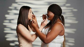 interracial belleza modelos dando cada otro cambios de imagen, utilizando hacer arriba cepillos y sensación alegre en estudio. joven amigos disfrutando divertido protección de la piel campaña, cuerpo positividad y aceptación. foto