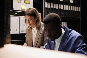 Diverse private investigators checking fraude case, analyzing police evidence late at night in arhive room. Detectives discussing crime scene, discovering files with confidential victim report photo