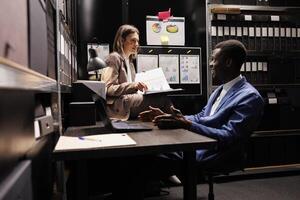 Diverse bookkeepers analyzing management files, working late at night at accountancy report in arhive room. Managers checking bureaucracy record, reading administrative documents photo