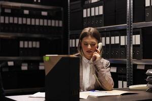 Bookkeeper discussing accountancy report with manager using landline phone, working overhours at management research in storage room. Repository worker checking bureaucracy record photo