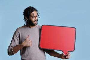 Smiling arab man holding empty red speech bubble and showing thumb up. Arabian person with happy facial expression standing with blank dialog frame for advertising message photo