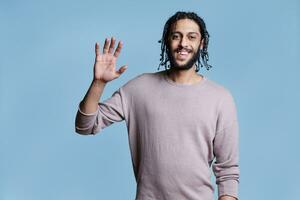 Friendly smiling arab man waving hello and looking at camera with positive emotions. Cheerful arabian handsome model wearing casual clothes raising hand for greeting portrait photo