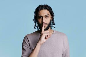 Arab man making quiet gesture and holding forefinger on lips portrait. Young person with serious facial expression doing tsss sign and looking at camera on blue background photo