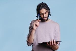 Pensive arab man thinking while holding clipboard and planning. Thoughtful young adult person in doubt reading checklist and touching face with pen with puzzled expression photo