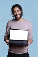 Cheerful arab man standing with laptop showing blank screen for web page mock up portrait. Person holding portable computer with white display for advertisement and looking at camera photo