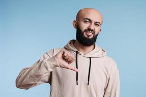 árabe hombre demostración disgusto con pulgar abajo, expresando negativo emoción retrato. joven persona con desaprobación cara mueca posando con desacuerdo firmar y mirando a cámara foto