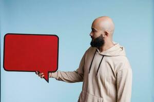 árabe hombre publicidad producto con vacío rojo bandera, demostración marco para texto. joven persona participación blanco diálogo burbuja forma con capa pluvial espacio para promocional mensaje burlarse de arriba foto