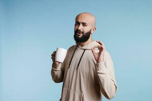Arab man enjoying coffee and showing ok gesture with fingers portrait. Young person drinking tea from white mug and looking at camera with approval sign and satisfaction facial expression photo