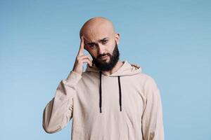 Arab man feeling pain of migraine while looking at camera with tired facial expression. Person rubbing temple while suffering from headache studio portrait on blue background photo