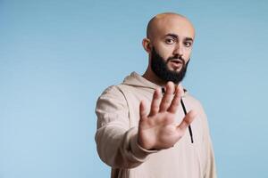 Young serious arab man showing prohibition gesture with palm portrait. Person making stop and rejection signal with hand while looking at camera with confident facial expression photo