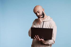 Thoughtful arab man rubbing chin in doubt while working on laptop. Young pensive person with puzzled expression thinking while holding portable computer and analyzing business data photo