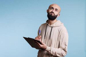 Thoughtful arab man holding clipboard and looking upwards with pensive facial expression. Puzzled person remembering while taking notes and writing list on blue background photo