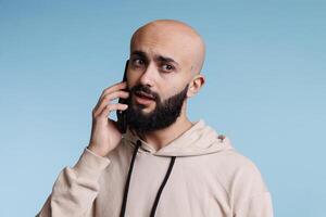 Unsure arab man talking on smartphone and looking away with uncertain expression. Young bald bearded person in doubt while answering mobile phone call and having communication photo