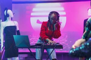 mujer DJ en auriculares mezcla sonido con controlador mientras ejecutando en etapa con focos en Club nocturno. africano americano músico utilizando digital música mezclador durante espectáculo en club foto