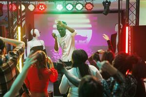 Crowd dancing and raising hands while musician performing on stage in club. People moving on dancefloor and having fun while enjoying live electronic music concert in nightclub photo