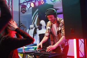 Dj in headphones using jog wheel and knobs while mixing sound with controller on nightclub stage. Young man musician playing electronic music during disco party in dark club photo