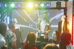 People dancing and enjoying dj show at disco party in nightclub. Young man musician performing and playing electronic music on stage for crowd in club illuminated with lights photo