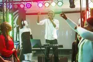 africano americano músico canto y jugando utilizando controlador en etapa en Club nocturno. hombre DJ con micrófono ejecutando a electrónico música concierto en club mientras multitud bailando foto