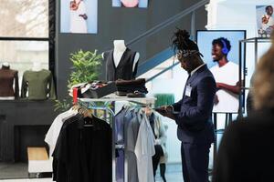 African american employee checking clothes inventory on tablet computer, looking at fashionable merchandise on hangers. Showroom worker checking discount list on device in shopping centre photo