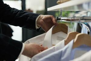 Caucasian man looking at hangers filled with merchandise in showroom, shopping for fashionable clothes. Senior customer buying casual wear to increase wardrobe in clothing store. Close up photo