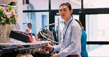 Asian worker arranging hangers with trendy clothes, working at clothing store visual. Cheerful manager preparing modern boutique store for opening, checking racks full with new fashion collection photo