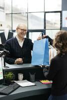 Senior client taking shopping bag from store worker, standing at counter desk paying for purchase in fashion boutique. Elderly man buying modern clothes making electronic transaction at mall photo